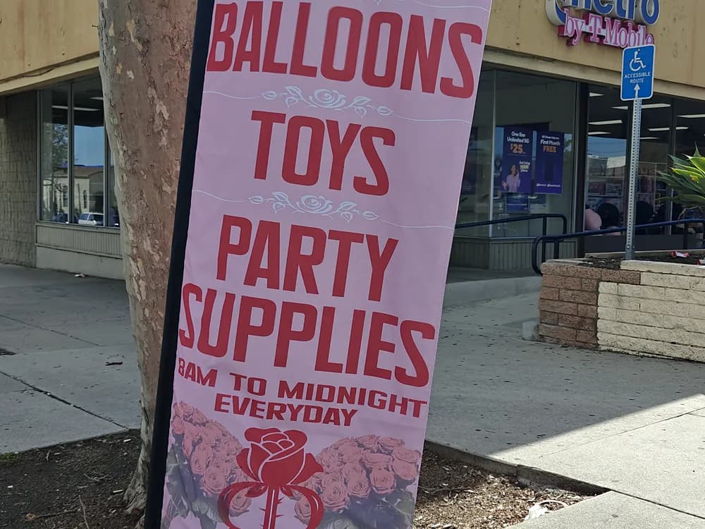 Tall printed vinyl banner reading Balloons Toys Party Supplies outside a retail unit