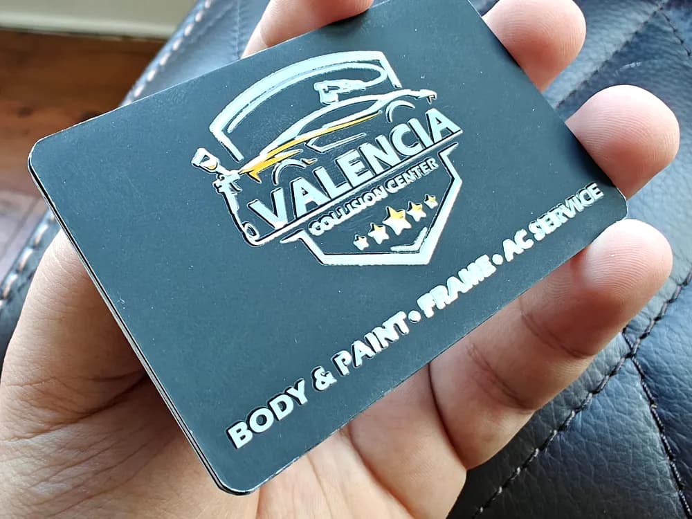 Teal soft-touch business card with a spot-gloss shield logo held in a hand