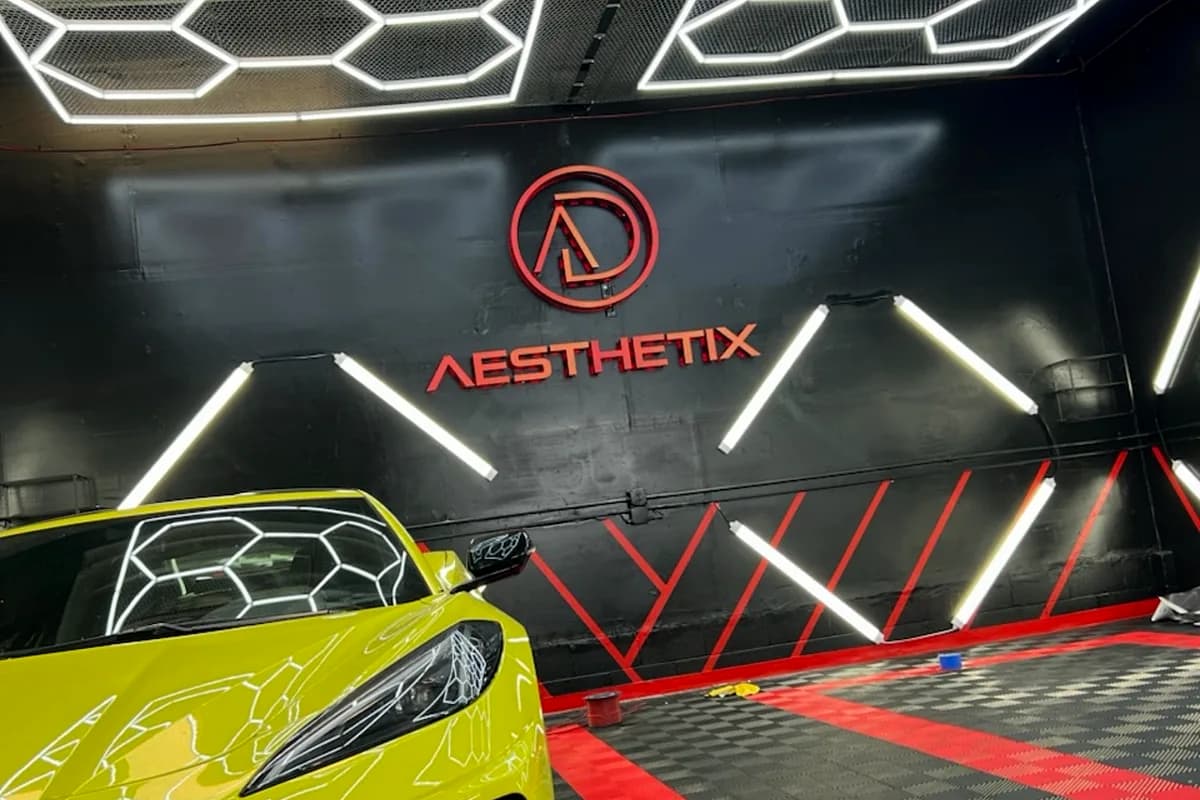 Backlit showroom wall logo above a yellow sports car in the Aesthetix detailing studio