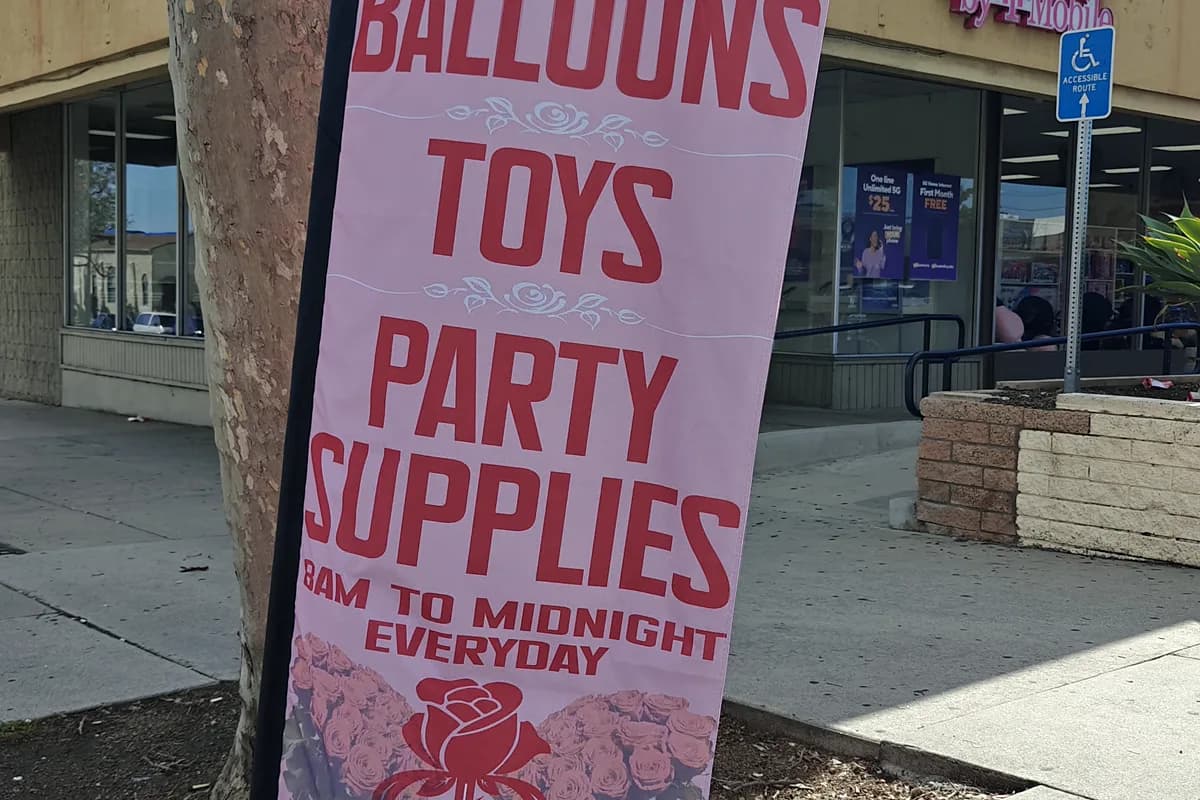 Tall printed storefront banner reading Balloons Toys Party Supplies outside a retail unit